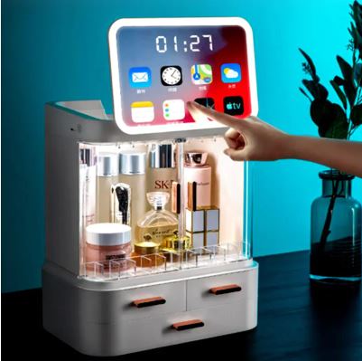 A hand touches the My Store Boîte de rangement à écran LED, displaying app icons and a clock, filled with beauty products and set on a table with a vase in the background.