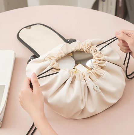 A person opens a beige Sac de maquillage à cordon by My Store on a light pink table, revealing cosmetics and personal items inside.