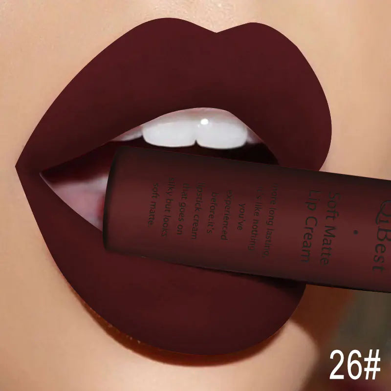 A close-up of lips with dark matte color holds a tube of Léa Beauty & Cosmetics Matte Non-Stick Liquid Lip Gloss—Long-Lasting Finish between the teeth. The stylized image highlights smooth skin and shade 26# is shown in the lower right corner.