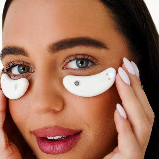 A woman with manicured nails holds two white My Store Capsules oculaires featuring small metallic accents under her eyes, looking directly at the camera.