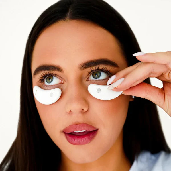 A woman with long dark hair and pink lipstick uses two My Store Capsules oculaires—holding one near her left eye and wearing the other below her right eye—for a soothing relief against a plain background.