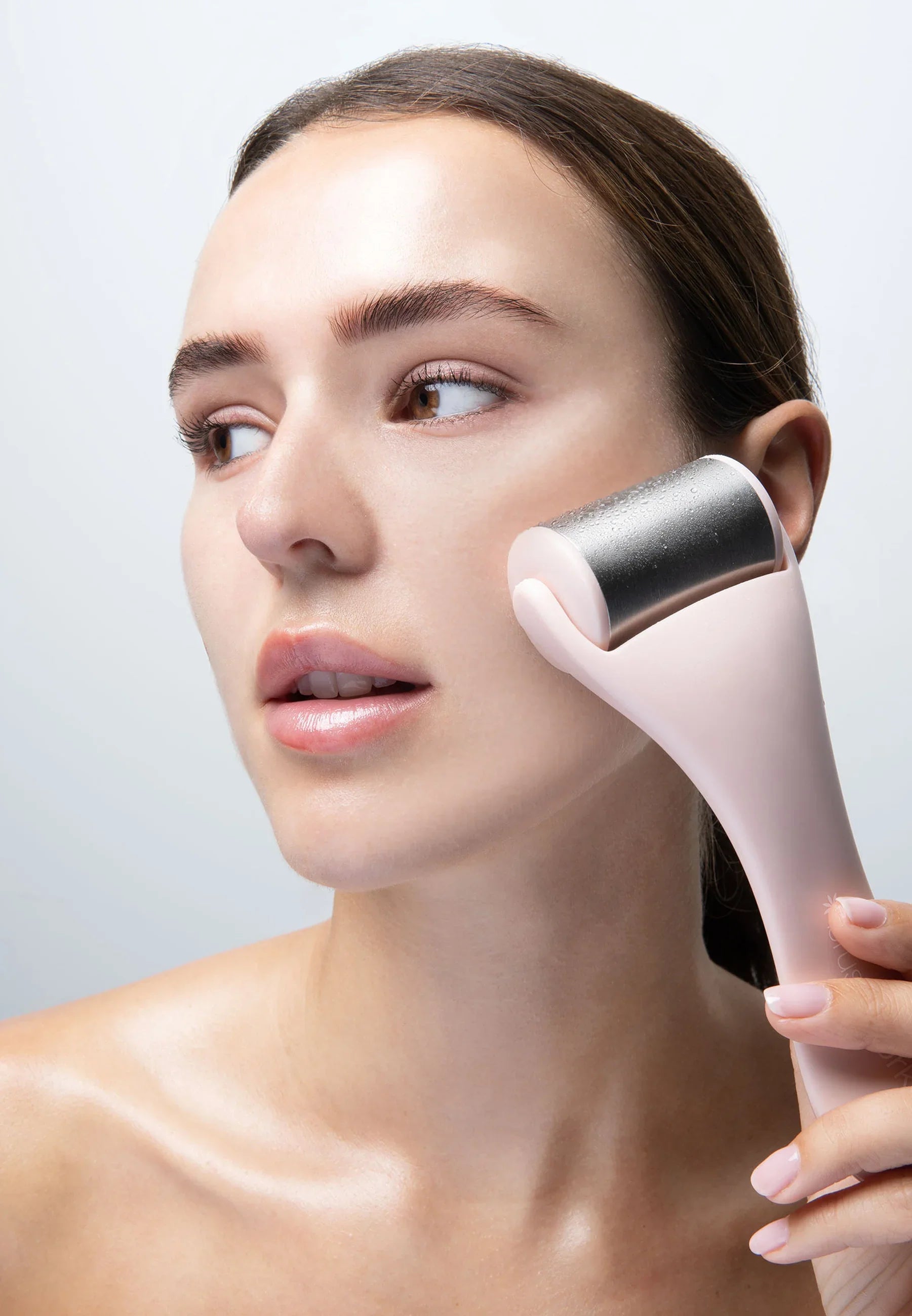 A woman glides the My Store Roulette de glace dermaroller with a pink handle across her cheek, enjoying its cooling sensation against a plain light background.