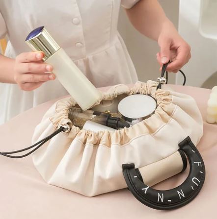 A person in white places a bottle into a beige My Store Sac de maquillage à cordon with black handles, surrounded by cosmetic items.