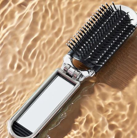 La Brosse à cheveux avec miroir pliant de voyage My Store, dotée de poils démêlants, repose sur une surface ondulée d’eau marron clair.